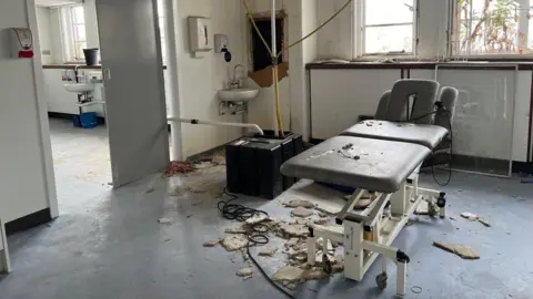 A hospital bed in the middle of a room surrounded by crumbling plaster.