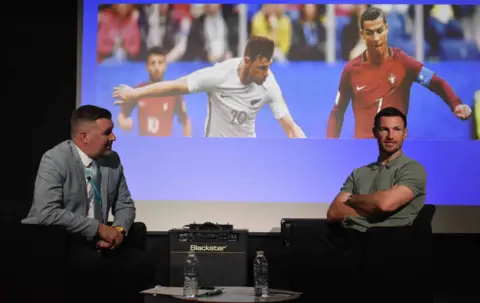 Suffolk New College Alan Pease on the left of the picture is seated and looking at Tommy Smith who is facing the audience, also seated. An image from his playing career is behind them on a screen