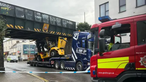 A side-on view showing the whole of the blue lorry and the yellow forklift on its back which is wedged against the bridge. In front of both is a parked-up fire engine with largely red livery with a yellow patch on its door