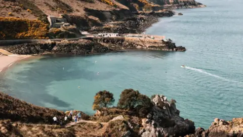 Matt Porteous A wide shot of a cove in Jersey. The rocky shore is surrounded by a light blue body of water.