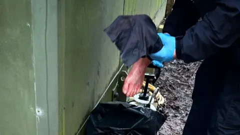 A police officer in dark blue overalls and wearing light blue latex gloves holds a replica of a human foot up to the camera.