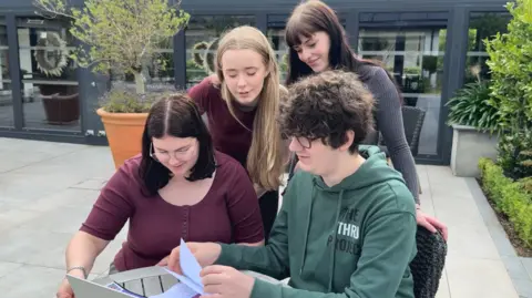 Four young people, two standing and two sitting, are looking at a laptop and some paper.