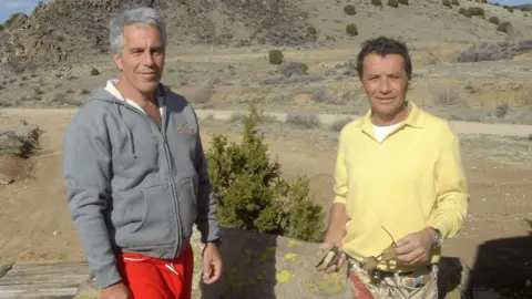 US Department of Justice Two men stand next to each other outdoors, smiling at the camera. The landscape behind them is dry with bits of scattered vegetation. Esptein has short grey hair and a grey hoodie. Brunel has short dark hair and a yellow top. He is holding a pair of sunglasses. 