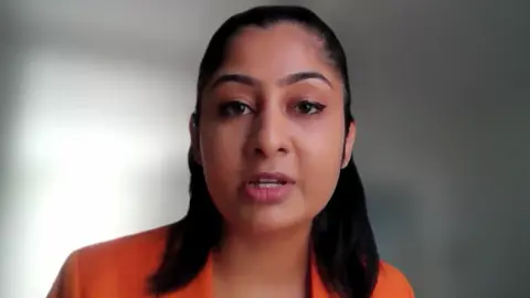 A woman wearing an orange jacket in a screenshot from an online video call. She has black shoulder-length hair.