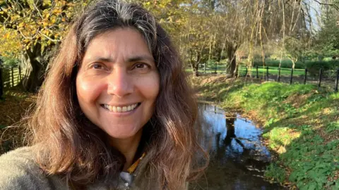 Dr Pallavi Devulapalli, former Green Party health spokesperson, standing outdoors near a small stream with autumn trees and a clear blue sky in the background.