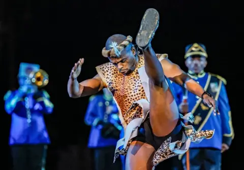 YURI KOCHETKOV/EPA Anggota South African Force Band tampil selama pembukaan Festival Musik Militer Internasional 'Menara Spasskaya' di Lapangan Merah di Moskow. Di latar depan gambar tersebut, seorang penari pria dengan bersemangat menendang kakinya tinggi-tinggi di udara, mengenakan pakaian dan hiasan kepala berbahan kulit macan tutul. Di latar belakang, terdapat anggota South African Force Band. Salah satu dari mereka memainkan terompet. Mereka mengenakan jaket biru dengan hiasan emas.