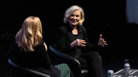 Glasgow Film Festival Jessica Lange on stage in Glasgow, wearing a dark suit and blouse. She is seated, facing the camera, talking to a chair woman who is turned away from us wearing a green and black spotted shirt and dark top. She has blonde hair. There is a table with water in front of them.