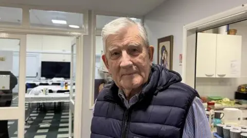 A man with short white hair, parted on one side, looks into the camera. He is clean shaven and wears a blue puffy waistcoat jacket over a striped blue shirt. He is in a room inside a building with an open doorway into a kitchen on one side and another doorway leads to a dining area.