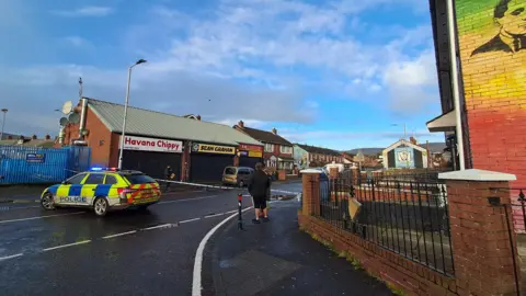 BBC A police car in the middle of the road which has been closed with tape. A set of shops can be seen on the other side of the road. 
