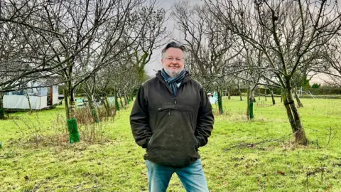 A man in a blue scarf, dark green coat and blue jeans stands in an orchard. Over twenty bare trees can be seen behind him. The man has glasses and fairly long dark hair. There is a shelter in the distance. The sky is cloudy.