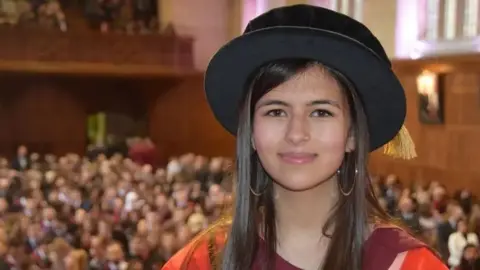 University of Bristol A young woman with long dark hair in PHD graduation robes stands in front of a hall full of people. the crowd behind her is out of focus.