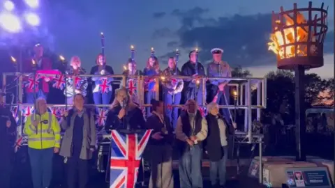 Christchurch Town Council Two rows of people holding torches and standing on a platform decorated with union flags. In front of them a woman with a microphone is singing. Her music stand is decorated with a large union flag.
