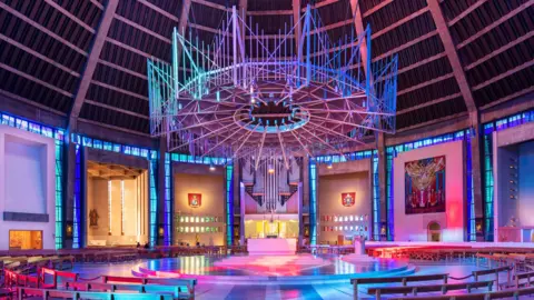 Historic England Archive A large circular chandelier-style feature with a hole in the centre hangs from the ceiling, above a circular altar. Rows of wooden benches encircle the stage and the cathedral is lit in blue, pink, purple, pink and orange.