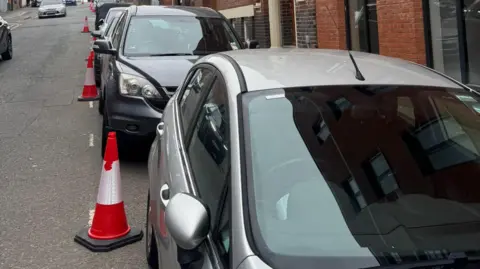 Joseph Butler Grey cars on a street in Birmingham's Jewellery Quarter, all the cars have Penalty Charge Notices on the windscreen. A row of orange, white and black traffic cones marks where parking restrictions were imposed.
