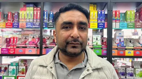 Javed Iqbal looking straight at the camera. He has dark hair. He is wearing a light-coloured jacket over a grey polo shirt. He is standing in front of several shelves containing vapes.