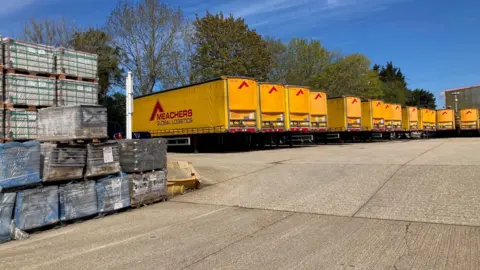 A line of yellow lorries with 'Meachers Global Logistics' printed on their side all sit in a yard. Next to them is a tall stack of wrapped pallets.