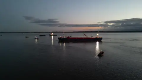 The vessel is afloat in the Humber, at dusk. The sun has set and the vessel and tug boats have their lights on.