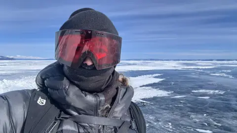 Anya Eames Anya poses in her cold-weather gear, with the snow covered frozen lake behind her. She is wearing a thick black coat, a black hat, a thick neck buff and large red-tinted goggles, and has a black backpack on. 