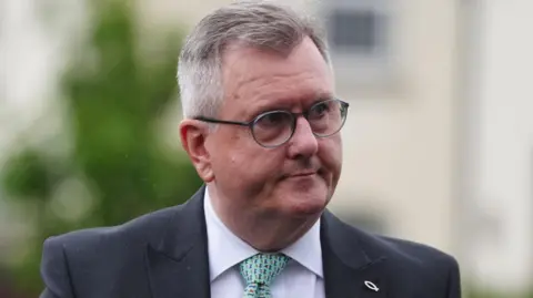 A man with short, cropped greying hair is seen in close up, with just his face and the tops of his shoulders visible. He is wearing dark-rimmed glasses, a dark blue suit jacket, white shirt and green patterned tie. 