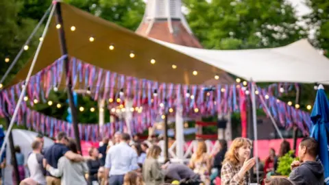 Mary Doggett A tent with pink and blue streamers and tassels which has been put up in Chapelfield Gardens. There are fairy lights on the tent and crowds of people.