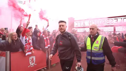 Reuters Middlesbrough manager Kim Hellberg arrives at the Riverside stadium before the match, escorted by someone in a high-vis jacket. Dozens of fans, wearing red and white, are stood behind barriers with some shouting. Others have their hands out as if to high-five Hellberg. Multiple red flares have been lit, leaving the scene with red dust around it.