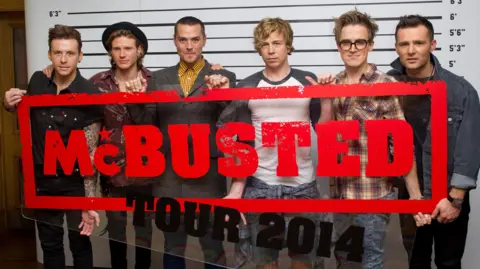 Getty Images McBusted holding a sign that says "McBusted Tour 2014". On the left is Danny Jones, on his right is Dougie Poynter, then Matt Willis, then James Bourne, then Tom Fletcher, then Harry Judd. They are all stood in front of a height chart.