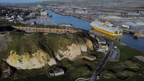 An aerial image of Newhaven Port - there are cliffs in the foreground, with a river to the right. There are buildings in the distance, and a white and yellow ferry on the river