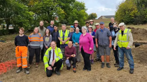Bedford Roman Villa Project A large group of people outside standing in a working archaeological site. Two are kneeling down, the rest are standing up. They are all smiling and looking at the camera. Trees are behind them, there is some orange netted fencing to the left and a house in the distance.