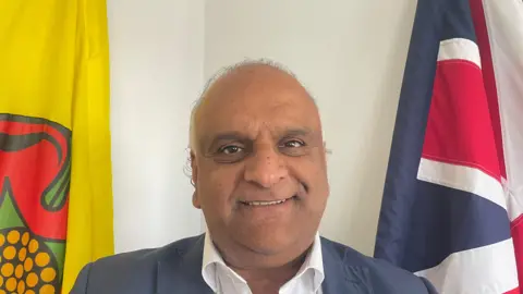An image of Councillor Azhar Ali. He's standing in front of a white wall, with the Lancashire flag to his left and the Union Flag to his right. He is smiling and wearing a navy suit and white shirt.
