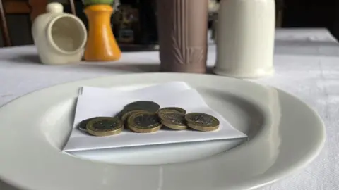 Andrew Turner/BBC Coins on a piece of white paper, on a plate at a table set with bottles and condiments - a picture representing a customer tip.