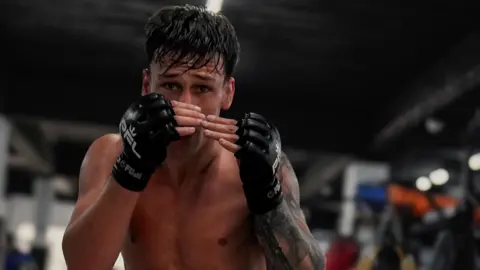 Liam Lens Liam McCracken is wearing protective MMA black gloves as he holds both hands up in a fighting stance. 