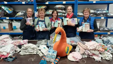 Five people holding boxes of baby bottle equipment. There are piles of baby clothes and a large stuffed orange stork on the table in front of them and further bags and boxes on blue warehouse shelves behind.