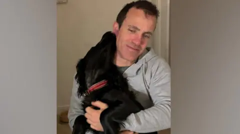Wiltshire Police Man smiling while cuddling a black dog indoors.