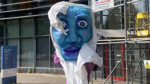 Bristol Experimental Puppet Festival A person holds a huge papier mache head outside a building covered in scaffolding. The head is painted blue. 