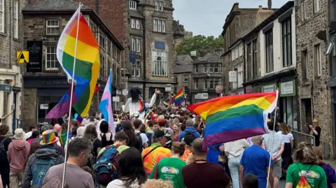 People fly rainbow pride flags as they stream through a historic narrow street in Lancaster city centre with old shops lining both sides of the road