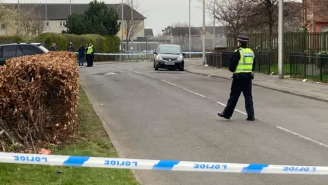 BBC Police at the scene of a shooting in Edinburgh. Police tape surrounds the scene, with officers walking about and speaking to residents