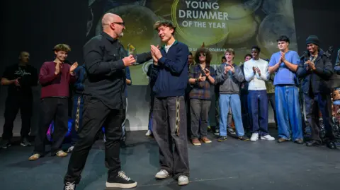 Dave Hughes/Avedis Zildjian Company Ellis Gates being handed the Young Drummer of the year award on stage. Other competitors stand behind him clapping