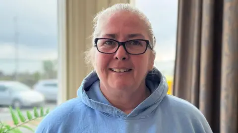 Shirley Cain is wearing a light blue hoodie. She has grey hair tied back and is wearing dark rimmed glasses. She's smiling and standing in a bay window with a fern plant to the left and a curtain to the right - there are parked cars are outside.