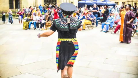 A woman in colourful, traditional African clothing performs in front of a seated crowd in a city square. It's a bright and sunny day. The performer's back is to the camera.