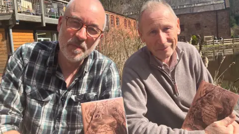 Two men are holding brown paper books while sat down looking at the camera.