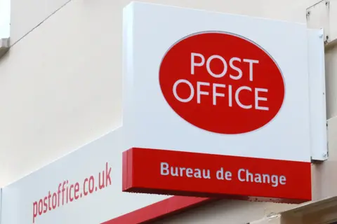 A white and red Post Office sign hangs from the side of a building. The words "Post Office" in white writing sit within a red circle. Below are the words "Bureau de Change", in similar white writing enclosed in a red rectangle.