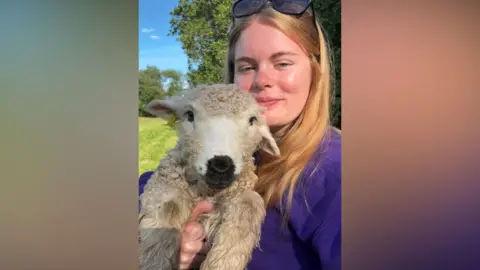 Jessica Tilley-Moore A young woman with blonde hair holds up a lamb and smiles at the camera.