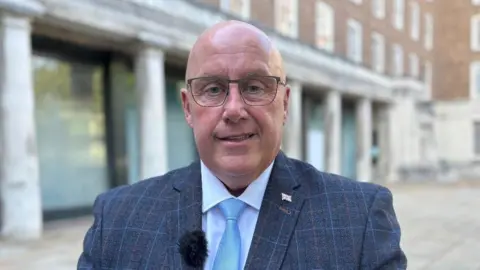 Mick Barton, Reform county council leader of Nottinghamshire, standing outside of County Hall wearing a blue, wool suit and light blue tie with a white shirt. He is a bald, middle-aged man wearing thin, metal-frames glasses. 
