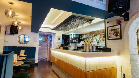 Greensmith Photography Inside the cocktail bar featuring blue velvet seats and cream and beige tiled bar filled with drinks and cocktail shakers.