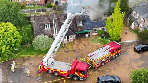 A fire engine and a ladder platform parked outside a house, surrounded by hoses. Two firefighters up the ladder look over the house as smoke billows from its roof.