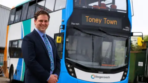 Stagecoach Midlands A man in a navy blue suit and blue shirt and tie stands in front of a double decker bus. "Toney Town" is written on the front in orange letters. 