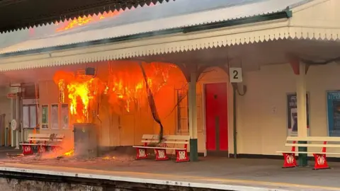 Devon and Somerset Fire and Rescue Service The image shows a railway station platform with part of the station building engulfed in a large fire. Bright orange flames are pouring out of the structure and through the roof, while a jet of water from firefighters can be seen spraying towards the blaze from the left-hand side, with railway tracks running across the foreground.
