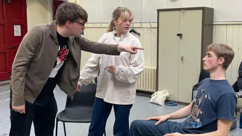 A rehearsal of a scene in the play, performed in one of the university's rehearsal rooms. A man is shouting and pointing at another man, who sits looking dejected. Between them, a woman tries to mediate.