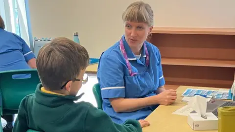 A nurse wearing a blue and white uniform with purple lanyard sits at a desk talking to a child who has their back to the camera. The pair are in an office with an empty shelf and another nurse visible in the background.