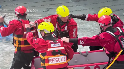 NIFRS A group of firemen in red and yellow hats. On their backs is 'RESCUE' written in red. They are standing in water and working together to move a white and red boat along.
